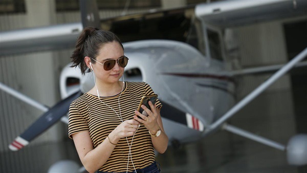 photoshoot of young woman standing near/cleaning an airplane listening to her phone via white Apple ear buds.