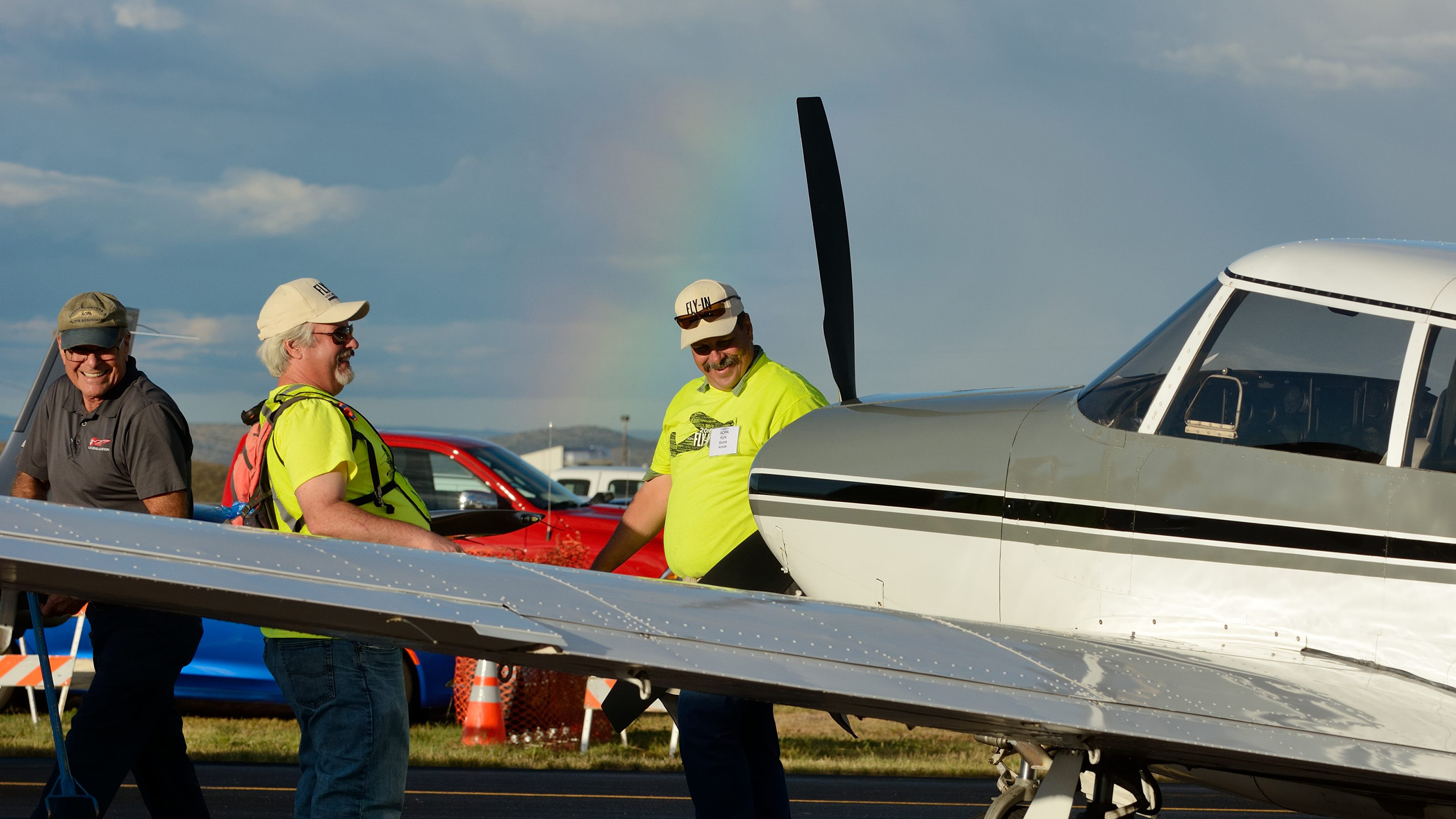 Airside volunteers share a laugh after positioning a Piper Comanche in the static display. A rainbow gives evidence of an afternoon rainshower. Photo by Mike Collins. Airside volunteers share a laugh after positioning a Piper Comanche in the static display. A rainbow gives evidence of an afternoon rainshower. Photo by Mike Collins.