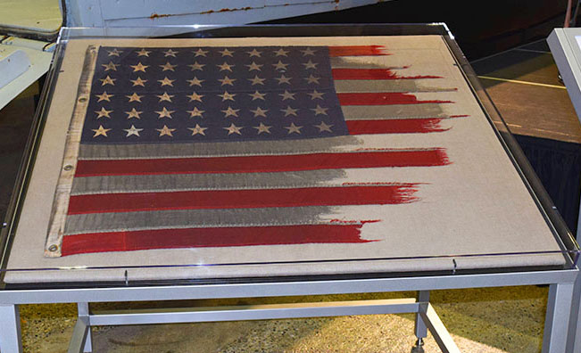 This flag, displayed at the Flying Heritage & Combat Armor Museum, was carried into combat on D-Day on LCT-595, a Landing Craft Tank amphibious assault vessel. LCT-595 and three other LCTs were some of the first vessels into combat that morning. LCT-595 delivered its American Army swimming tanks under heavy German machine gun fire. Another LCT was destroyed by a German mine. After the attack, LCT-595’s captain told US Navy Boatswain George Edward Rudisill to remove the tattered flag and fly a new full one. Rudisill asked to keep the battle-worn remains of the original D-Day flag. He preserved keepsake in a shoebox, along with the only known photograph of LCT-595. Photo courtesy FH&CAM.
