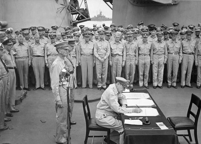 Gen. Douglas MacArthur signs formal surrender documents aboard the <em>USS Missouri</em> during ceremonies in Tokyo Bay on Sept. 2, 1945. Behind MacArthur are Lt. Gen. Jonathan Wainwright and Lt. Gen. A. E. Percival. Photo No. 280-G-348366 courtesy National Archives.